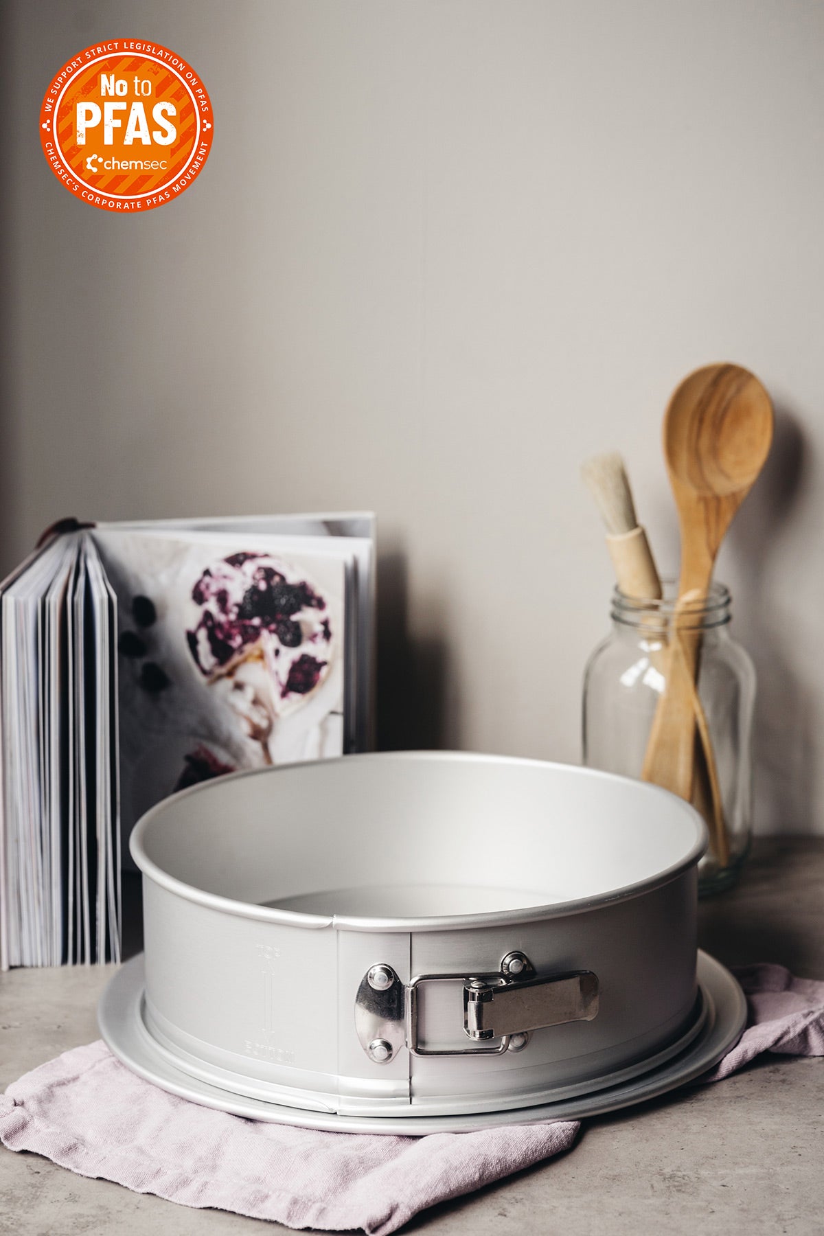 Anodisoidusta alumiinista valmistettu irtopohjavuoka valumareunalla aseteltu keittiön harmaan kivitason ja liilan pellavaliinan päälle.